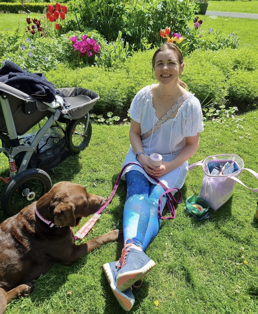 Image of Sarah sitting on the grass smiling with her assistance dog Millie beside her