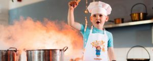 Young boy wearing a chef's hat waving a spoon over a bubbling pot on a stove.