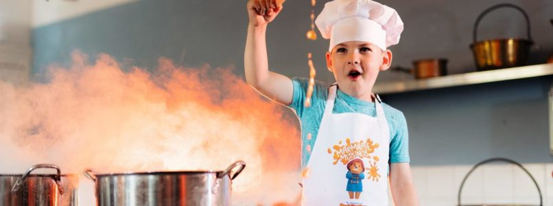 Young boy wearing a chef's hat waving a spoon over a bubbling pot on a stove.