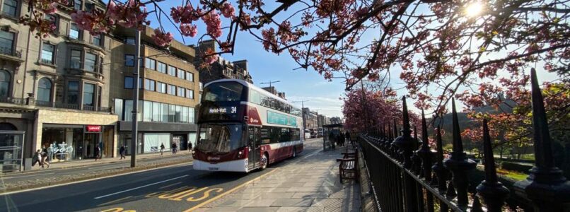 A Lothian city bus drives along Princes Street on a sunny day with blossom trees along the pavement.